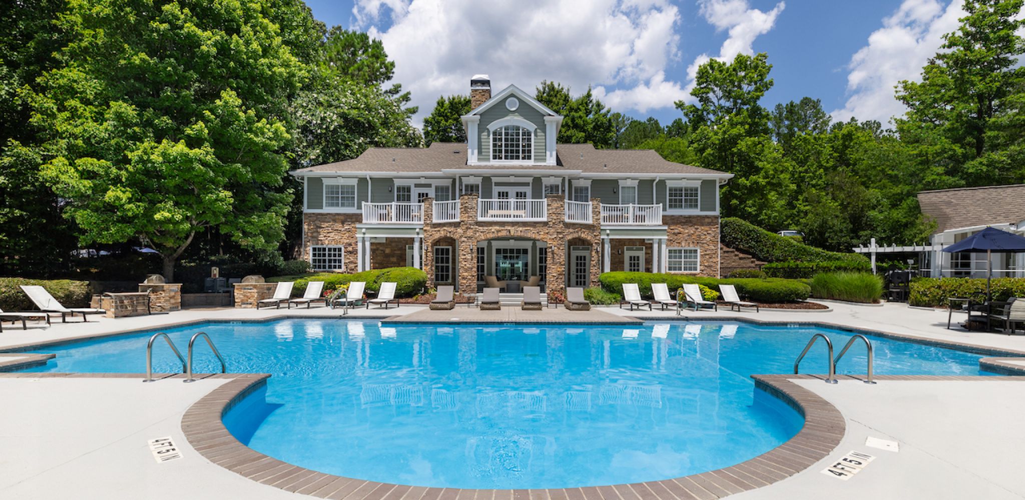 Hawthorne at Lake Heather luxurious pool area featuring a large, pristine pool surrounded by lounge chairs and lush greenery, with a beautiful two-story clubhouse in the background.