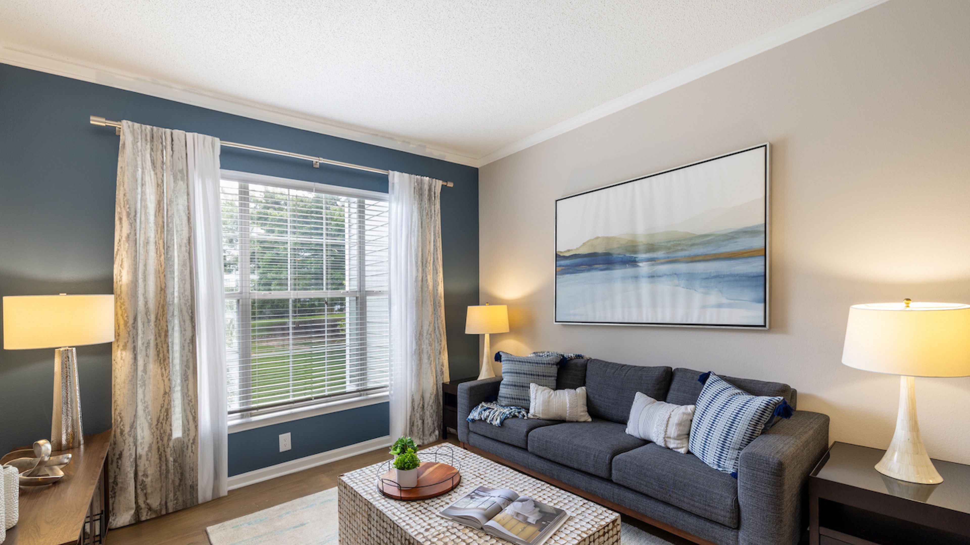 Hawthorne at Lake Heather stylish living room featuring a comfortable gray sofa, a textured coffee table, and large windows with elegant curtains that let in plenty of natural light.